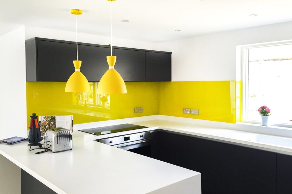 A yellow painted glass splasback in a kitchen with contrasting black cabinets and white worktops.