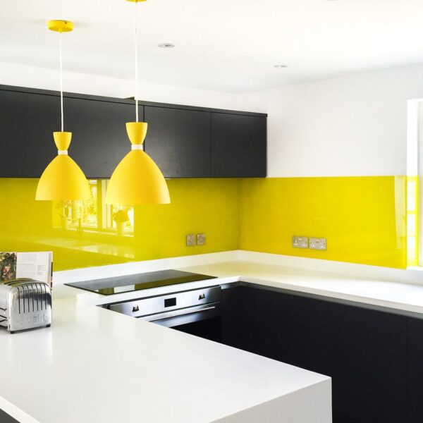 A yellow painted glass splasback in a kitchen with contrasting black cabinets and white worktops.